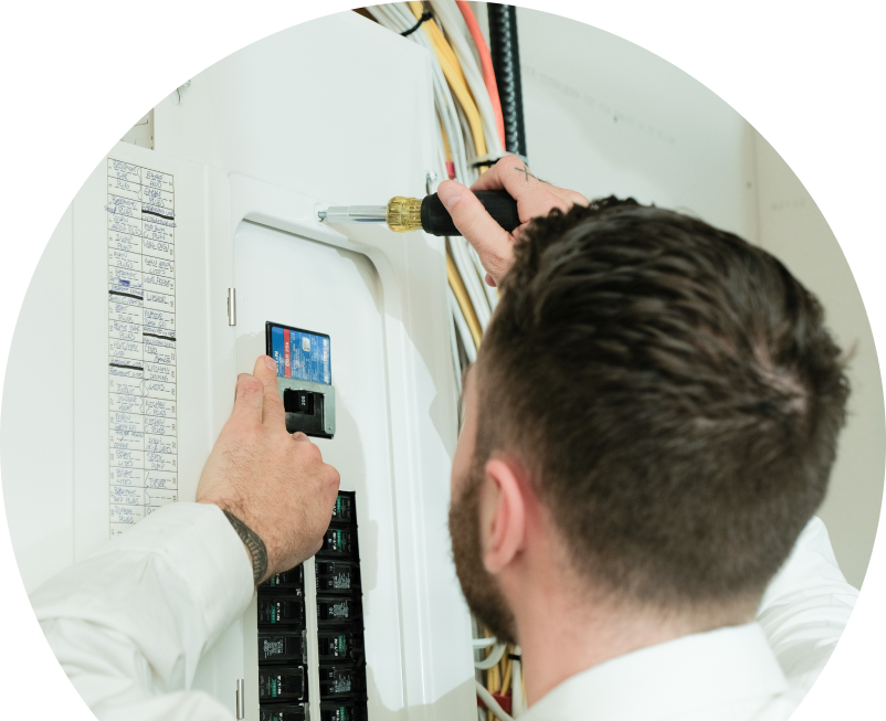 Wardlaw electrician using a screwdriver to perform an electrical panel upgrade, working near organized wiring in a residential wall.