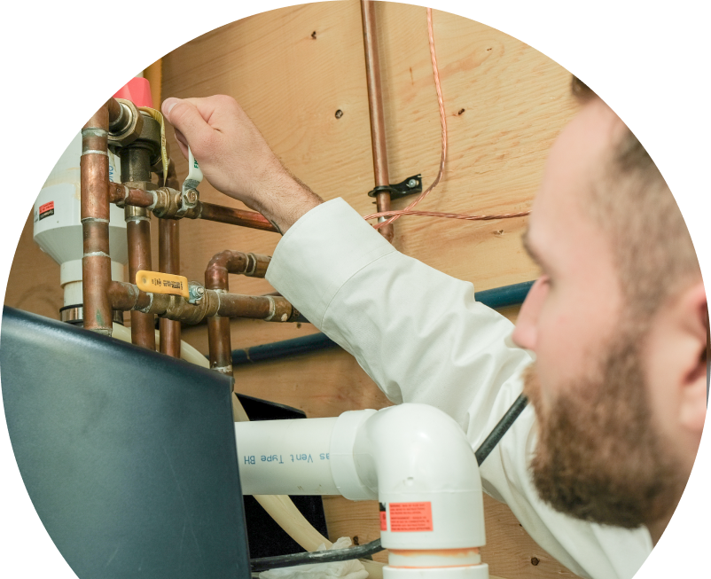 Wardlaw water heater specialist tightens a valve on copper piping during a water heater installation, with exposed wood paneling and wiring in the background. Wardlaw water heater specialist tightens a valve on copper piping during a water heater installation, with exposed wood paneling and wiring in the background.